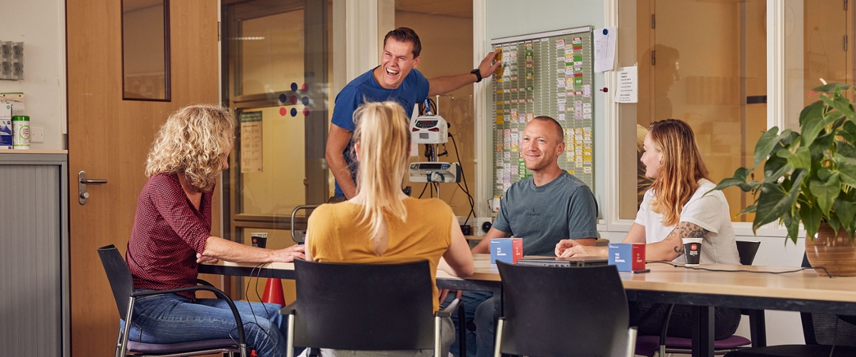 Vier leden van het psychiatrieteam zitten aan een vergadertafel. Een vijfde persoon staat naast de tafel en wijst wat aan op een bord.